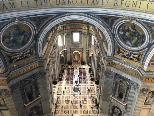 We are inside a colossal building, looking down to the floor far below — perhaps 150′ or more.  The structure appears to be pale marble, elaborately decorated and carved.  At the top of the picture, the far wall curves slightly towards us, suggesting we are at the opposite side of a round open space, perhaps the base of a dome; two massive pillars reach down from the corners of the photo to the floor below.  The curved section is colored gold, with tall blue lettering:  “…eam et tibi dabo claves regni c…”, etc.  The pillars have religious images, angels flanking, with crossed keys below, and more lettering below that.  The floor is decorated with mosaic patterns, and looking more closely, we can see dozens of people, tiny from our perspective.  We can see down into the choir, at least a hundred feet long.
