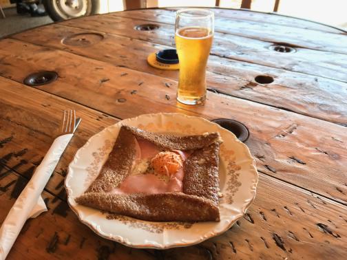 A buckwheat crêpe sits on a plate, partially folded around a slice of ham and a barely-cooked egg, with cheese scattered on top.  A small glass of cider is nearby.  The table is a large wooden spool that might once have been used for rope.