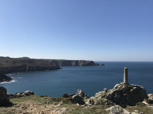 A small pillar, perhaps a monument, stands on a rocky outcropping at the edge of a nearby cliff.  Below is the Atlantic ocean, looking west towards more cliffs and a headland in the distance.