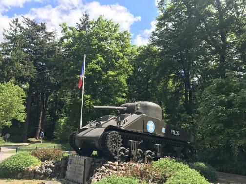 A stubby World War II–era tank stands to the east on a small rise, pointing left, as a monument.  It is painted olive green, with a light blue Free France sign and the word “VALOIS” in white.  Around it are some neatly-tended bushes, and a French flag.  In the background are tall trees, a lawn, and a sign for parking.  The small amount of visible sky is partly-cloudy.
