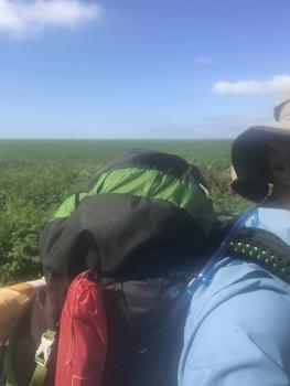 This selfie shows my backpack from my right side.  It is black and green; a tall, narrow red sack is on the side.  A blue tube, for water, snakes from the top of the backpack and over my shoulder.  Strapped to the back of the backpack is a loaf of bread.  All we can see of me is a blue sunshirt and a beige sun hat.