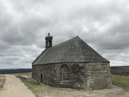 A squat stone chapel with a rounded end sits to the northwest on a flat, elevated site.  The chapel has an angled roof and a short bell tower, a small doorway, and a slightly larger stained-glass window.  To the left is a wide, beaten-down dirt path.  The sky is overcast.