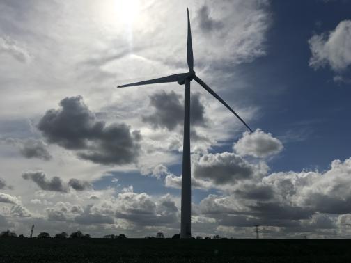 A massive éolienne (wind turbine) is silhouetted by the sun behind to the south-southwest.  The sky is partly cloudy; the field can’t be made out, though trees and power lines are visible on the horizon.