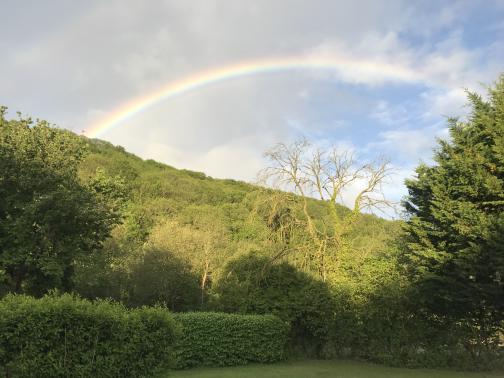 To the southwest, a steep, tree-covered hill rises to the left, with a bright rainbow visible in the partly-cloudy sky.  Nearer are a few individual trees, and a pair of hedges by a neat lawn.