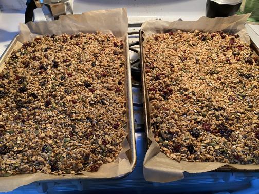 Two parchment paper–lined half-sheets of granola are cooling on the stovetop.  Identifiable are rolled oats, pumpkin seeds, sunflower seeds (shelled), dried cherries, and dried cranberries.